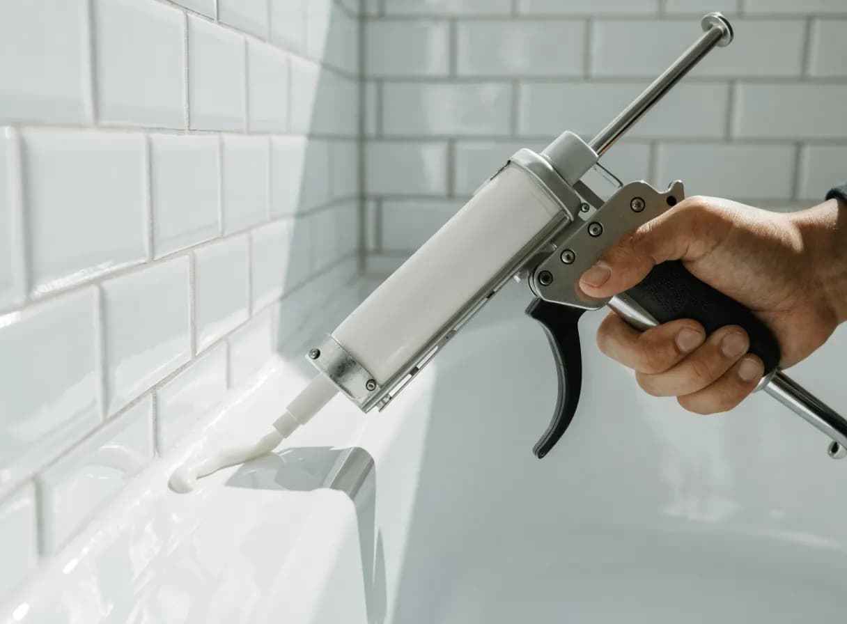 Caulk gun applying a clean white silicone bead where a bathtub meets white subway tile